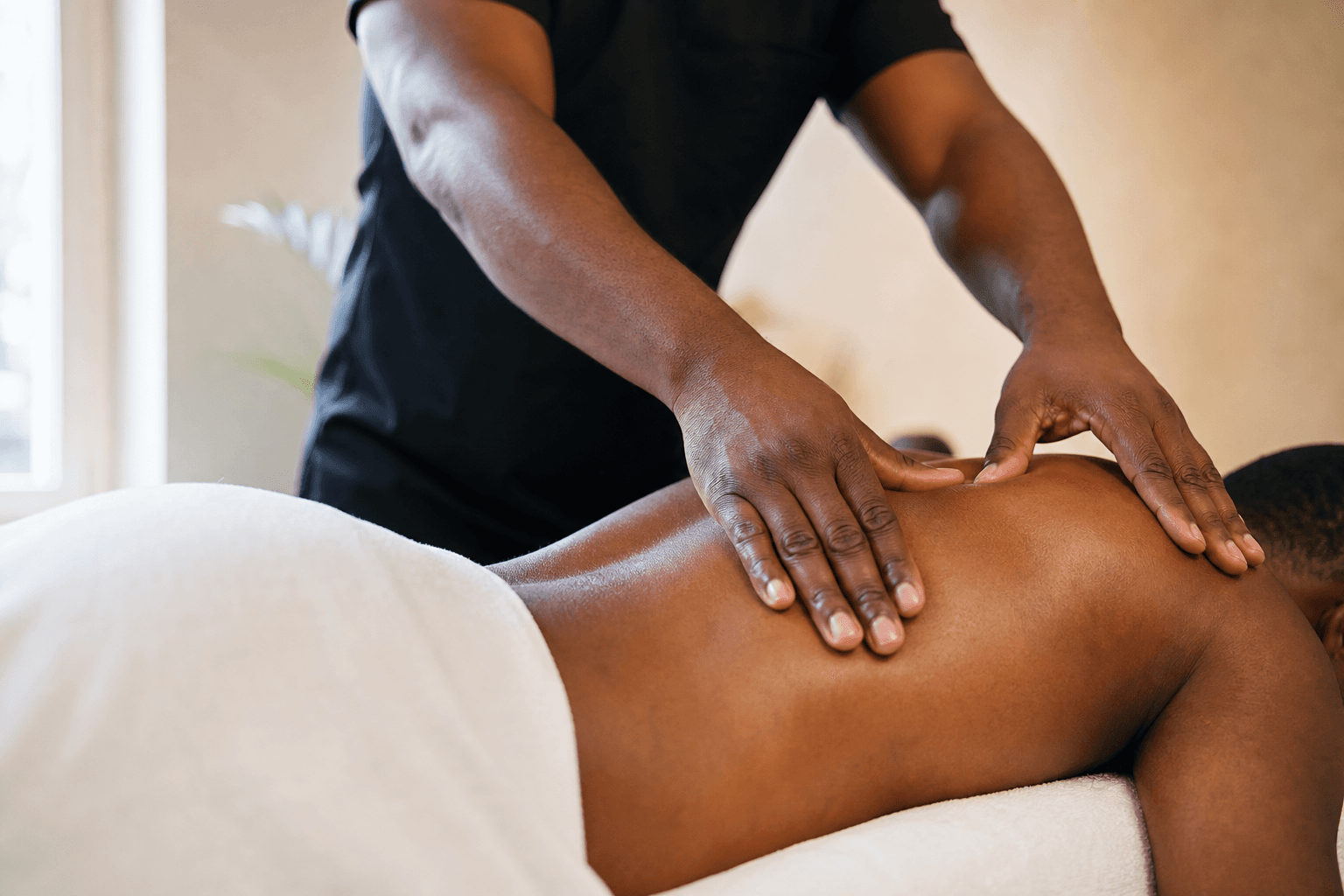 Black massage therapist's hands applying firm Swedish massage strokes across draped shoulders